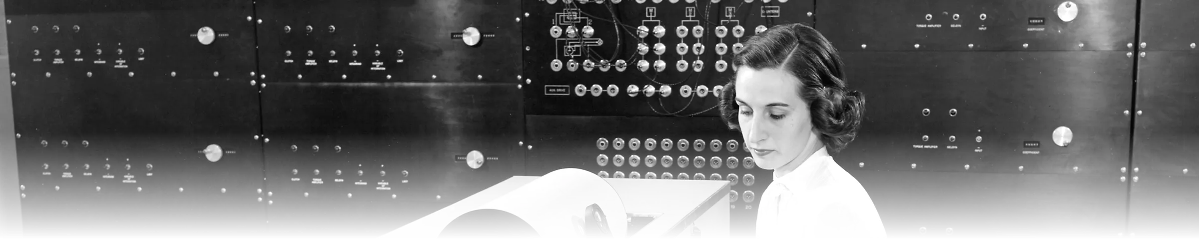 A black-and-white photograph of a technician operating the control console of a mechanical differential analyzer, in front of a dense wall of patch-panel switchboards