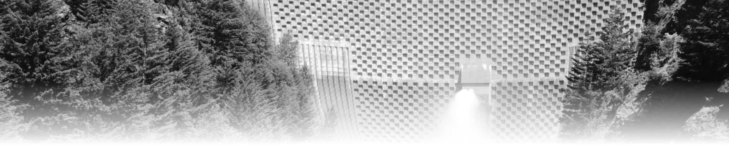 A black-and-white photograph of a dam with a relief valve open, water bursting out under pressure, flanked by dense conifer forest