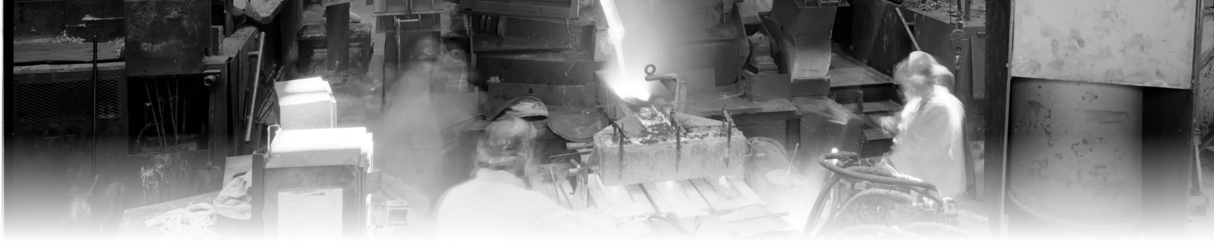 A black-and-white photograph of a foundry casting crew pouring molten metal from a holding furnace into molds, with a bright glowing pour stream lighting the scene
