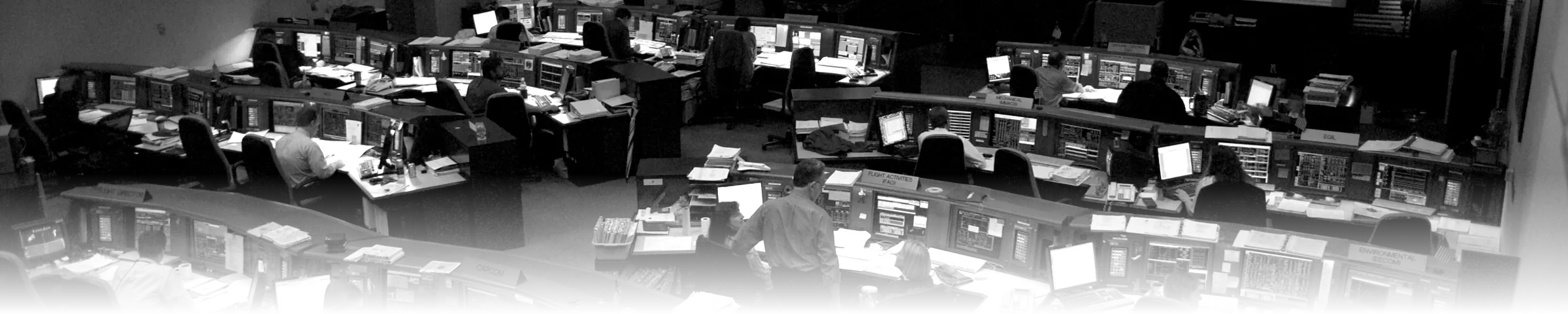 An elevated, black-and-white photograph of NASA Mission Control, showing rows of identically equipped console workstations