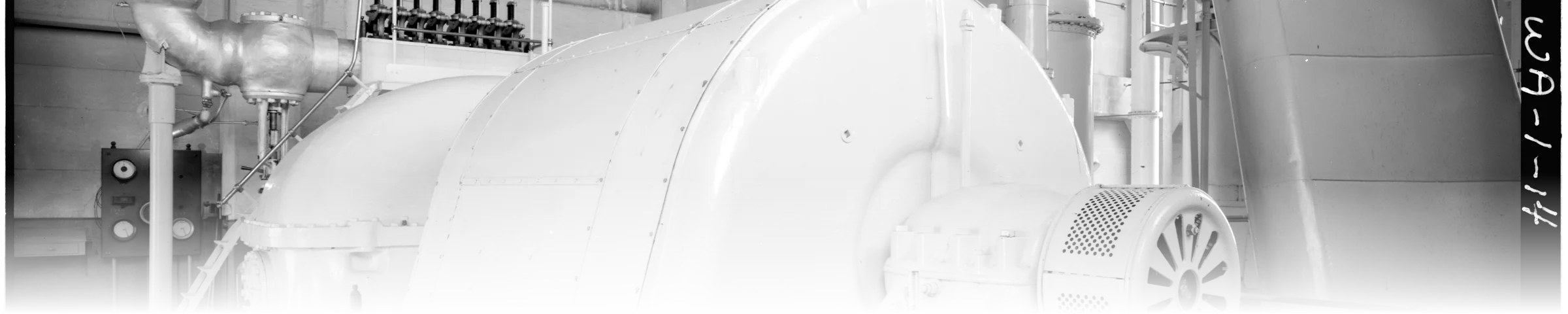 A black-and-white photograph of a large vertical steam turbine-generator unit, dense with piping, gauges, and a motor coupling