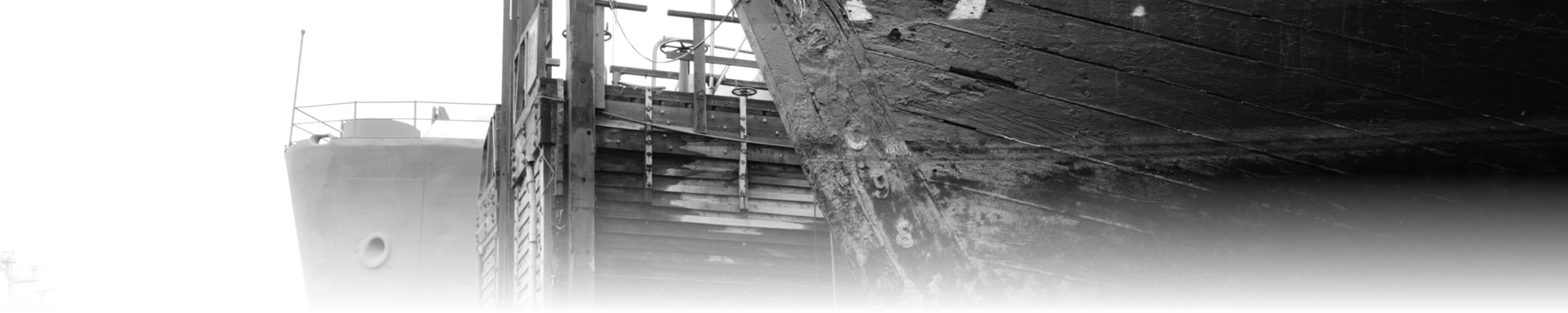 A black-and-white photograph of a steel reinforcement plate bolted onto the weathered wooden bow of a historic schooner in dry dock, with a second vessel and the dry dock wooden staging structure visible behind it