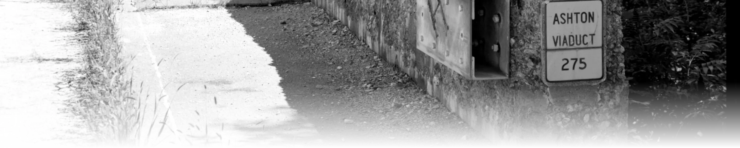 A black-and-white photograph of a cast metal "ASHTON VIADUCT 275" identification plaque bolted to a weathered stone bridge pier, alongside a utility box