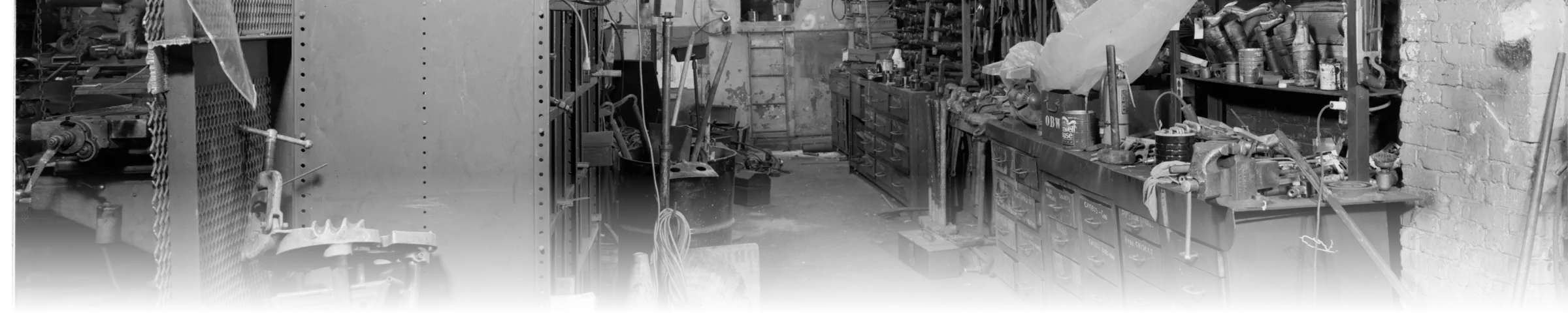 A black-and-white photograph of a machine shop tool room, with a mesh-fronted tool cabinet, a rack of hanging clamps and wrenches, and a run of individually labelled parts drawers under a workbench with a mounted vice