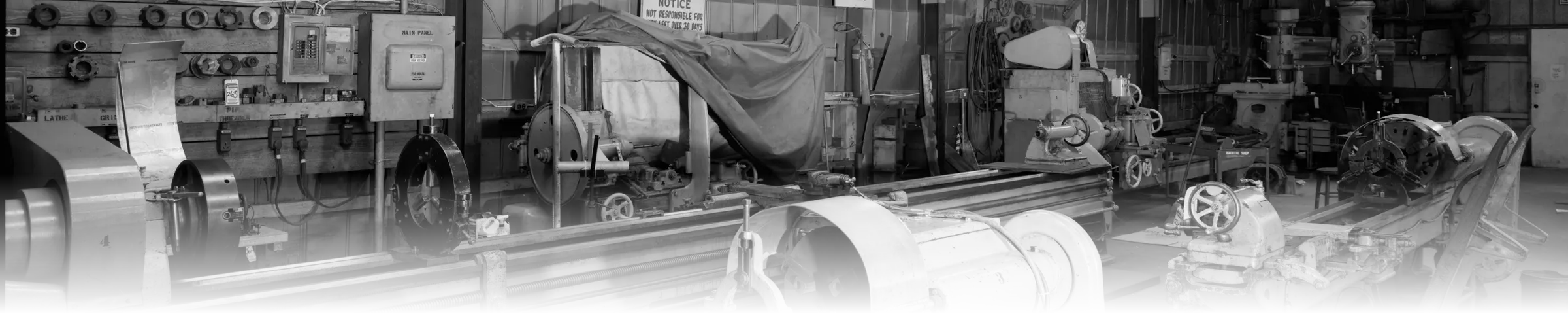 A black-and-white photograph of a machine shop interior, showing a wall of tool racks and chucks, an electrical panel, and several lathes